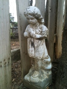 Statue of a little girl clasping her hands and looking down, next to a white picket fence